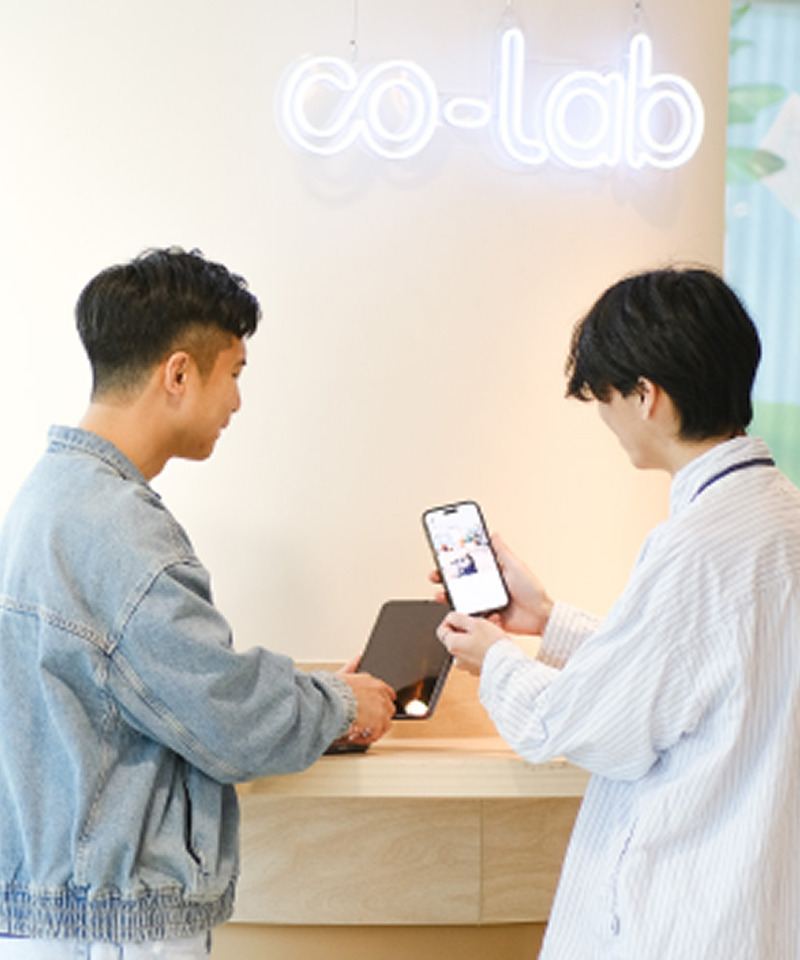 Two young people are interacting in a workspace called "co-lab." One of the boys in a denim jacket stands by the table, while another boy in a loose-fitting shirt is checking something on his phone.