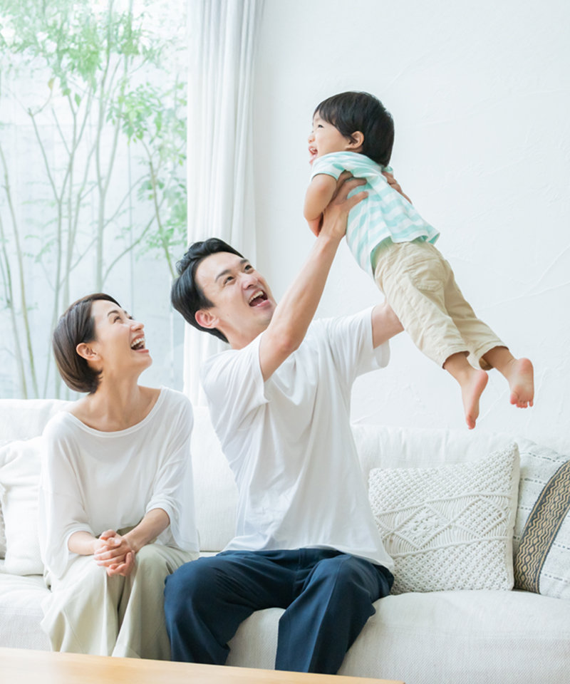 A couple sitting on a sofa, with the father lifting their child high, while the mother smiles at them, showcasing a joyful and warm family moment.​