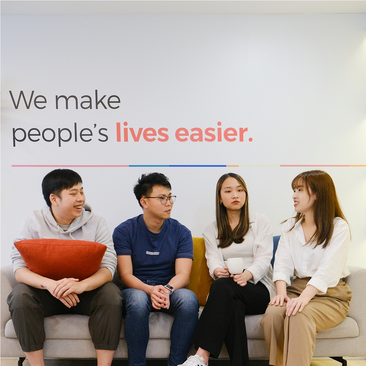 Four people sitting on a couch in front of a wall with the text “We make people‘s lives easier.” They are engaged in conversation, with a mix of expressions.