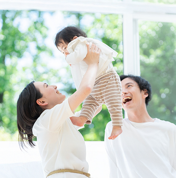A couple in a bright indoor setting, with the mother lifting their baby high, while the father smiles at them, showcasing a warm family atmosphere.​