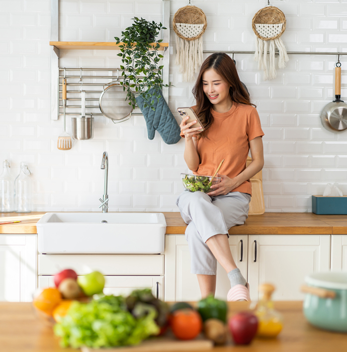 A woman sitting in a kitchen, holding a smartphone, with fresh vegetables and fruits nearby, showcasing a healthy lifestyle.​