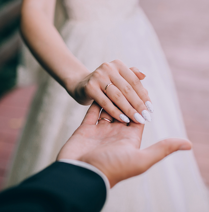 A close-up of a bride's hand holding the hand of a groom, showcasing a wedding band and elegant nails​​